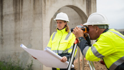 The survey team is using a theodolite and road construction plans. Civil engineers are measuring...