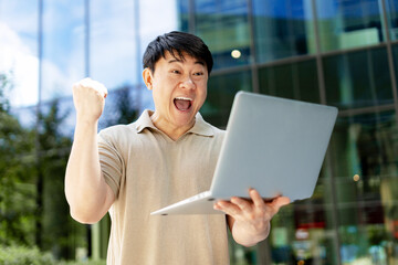 Excited businessman celebrating success while holding laptop in front of office building