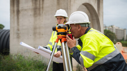 The survey team is using a theodolite and road construction plans. Civil engineers are measuring...