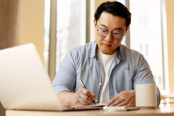 Asian Freelancer Taking Notes While Working on Laptop in Coworking Space