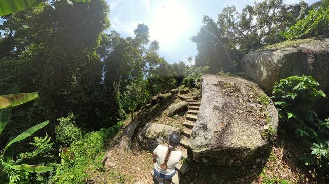 Woman Climbing Over Sunlit Rock Terrain Surrounded by Dense Jungle and Trees with Bright Blue Sky Above, Wide Angle View, Adventure Hiking Concept