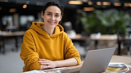 Attractive woman with long brown hair smiles while working on her laptop among color swatches and vibrant artwork in a sunny room