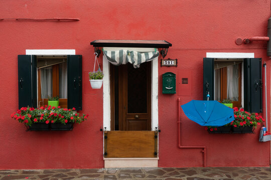 View of a charming red building with dark green shutters framing flower-filled windows, complemented by a turquoise umbrella, Venice, Veneto, Italy.