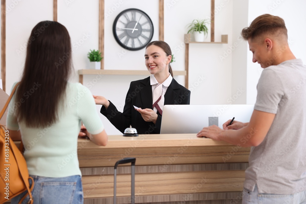 Poster smiling receptionist in uniform with hotel key card working with clients at reception desk indoors