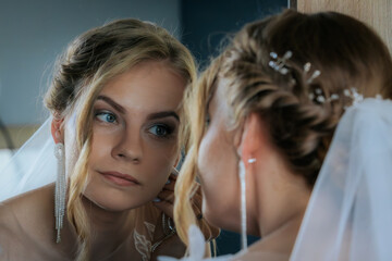 Bride looks into a mirror while adjusting her earring, wearing a lace gown and veil. A quiet, intimate moment of preparation before the ceremony.