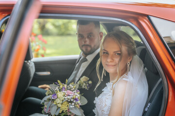 Bride and groom seated in the back of a decorated wedding car, smiling and holding a colorful bouquet. Natural light and greenery in the background.
