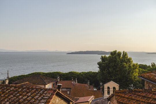 View of tiled rooftops cascade towards the tranquil lake and distant island under a serene sky in Passignano sul Trasimeno, Umbria, Italy.