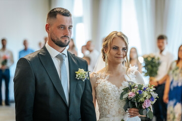 The bride and groom face a wedding officiant during their indoor ceremony, dressed formally and...