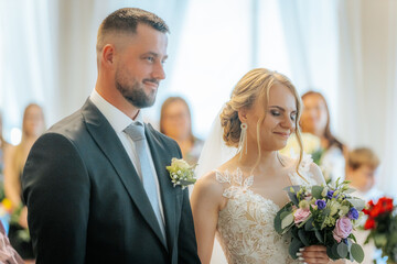 The bride and groom face a wedding officiant during their indoor ceremony, dressed formally and...