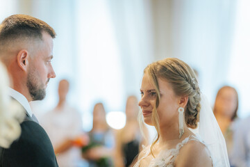 The bride and groom look into each others eyes during their indoor wedding ceremony, surrounded by softly focused guests in the background.