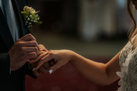 The groom gently places a wedding ring on the brides finger during their indoor ceremony, standing on a petal strewn red carpet aisle.