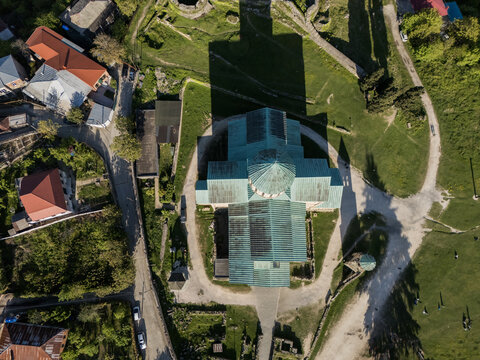 Aerial view of Bagrati Cathedral casting a long shadow, its turquoise roof contrasting with the surrounding green hills and quaint rooftops, Kutaisi, Imereti, Georgia.