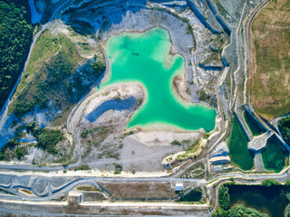 Aerial view of a vibrant turquoise lake nestled within the stark, grey landscape of a quarry, contrasting sharply with the surrounding green trees, Valkenburg, Limburg, Netherlands.