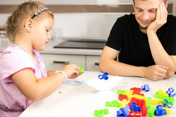 Young girl playing with colorful building blocks at a kitchen table while an adult watches, showcasing creativity and interaction in a playful learning environment