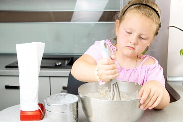 Young girl with blonde hair in pink shirt is mixing batter in a stainless steel bowl, surrounded by...