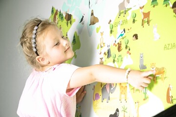 Young girl with curly hair in pink shirt joyfully points at colorful animal map on wall, engaging...