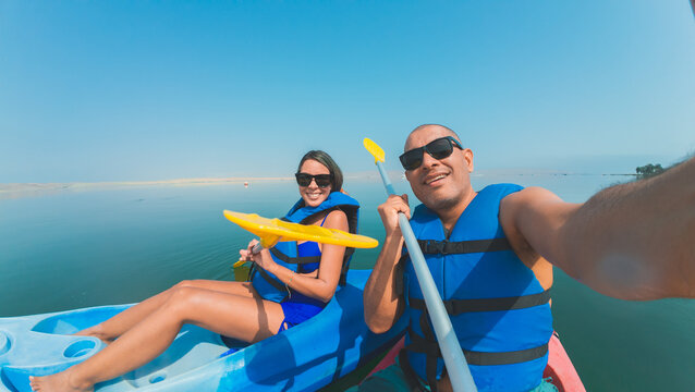 A man and woman are kayaking on a lake, wearing life jackets and holding paddles. The man is taking a selfie with his phone