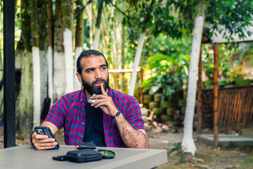 Man seated at table outdoors, raising an eyebrow while contemplating a decision with credit card...
