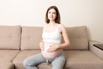 a pregnant girl sits at home on the bed and smears an anti-stretch mark cream on her stomach....