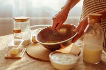 A person's hands are shown whisking ingredients in a metal bowl, with other baking essentials like butter, dry goods, and liquids in glass containers visible on a rustic table in warm light.