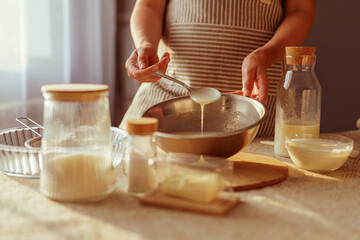 A person in an apron is adding batter from a spoon into a metal bowl, with various baking ingredients and utensils visible on a sunlit kitchen table.