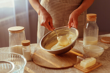 A person in a striped apron is stirring creamy batter in a metal bowl with a spoon, surrounded by various baking ingredients like butter and dry goods on a rustic table in warm natural light.