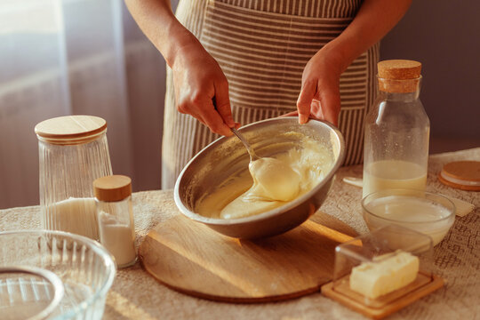 A person in a striped apron is stirring creamy batter in a metal bowl with a spoon, surrounded by various baking ingredients like butter and dry goods on a rustic table in warm natural light. - Powered by Adobe
