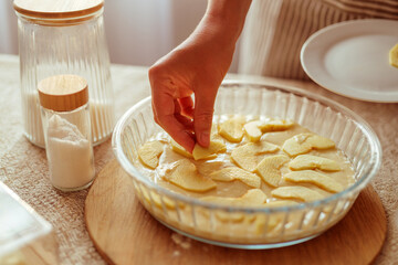 A person's hand is arranging sliced apples on top of batter in a glass pie dish, with a jar of dry ingredients in the background on a sunlit kitchen table.