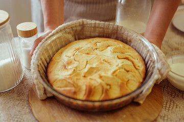 A person's hands are holding a freshly baked golden apple pie in a glass dish, wrapped in a cloth, resting on a wooden board, with other kitchen items blurred in the background.