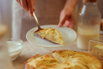 A person is serving a slice of freshly baked apple pie onto a white plate with a pie server, with the rest of the pie and other kitchen items blurred in the foreground and background.