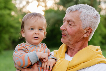 A loving outdoor portrait of a senior man with grey hair holding a baby with blue eyes and light brown hair, both looking to the side against a blurred green background with natural light.