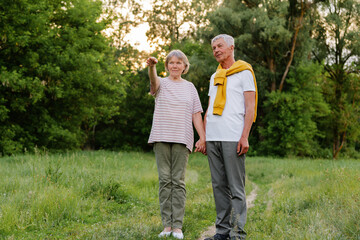A senior couple stands hand-in-hand in a grassy field, with the woman pointing into the distance while the man looks in the same direction, surrounded by lush green trees under natural light.