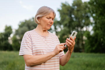 A thoughtful middle-aged woman with light, short hair is seen outdoors, looking down and interacting with a smartphone, with a blurred green natural background.