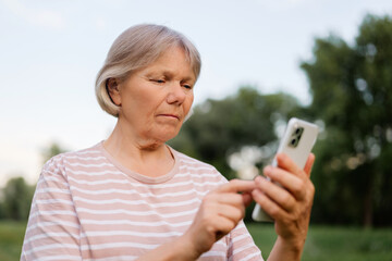 A middle-aged woman with light, short hair stands outdoors, intently focused on interacting with her smartphone, with a blurred green natural background.
