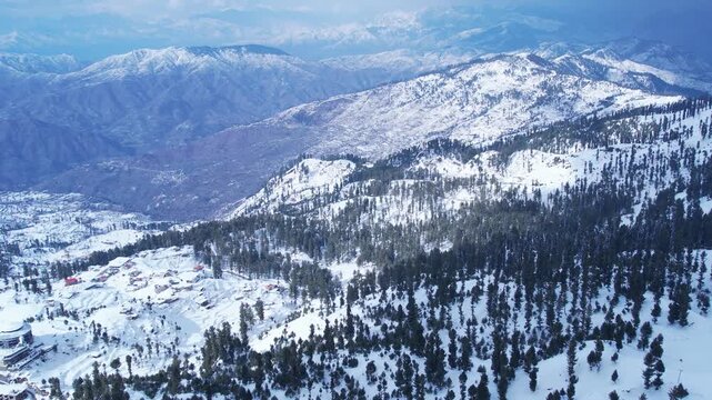 Sweeping aerial shot of a majestic snow-covered mountain range, with a village and hotel nestled in the vast winter landscape. Malam Jabba hill station.