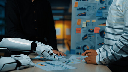 A human and a robotic arm engage in a collaborative discussion in a modern office, analyzing data charts and visuals to enhance productivity and innovation.Noogenesis