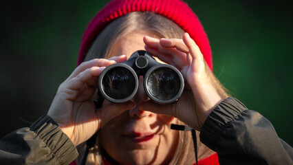 A girl with binoculars in her hands looks directly into the camera, wearing a red hat and a green...