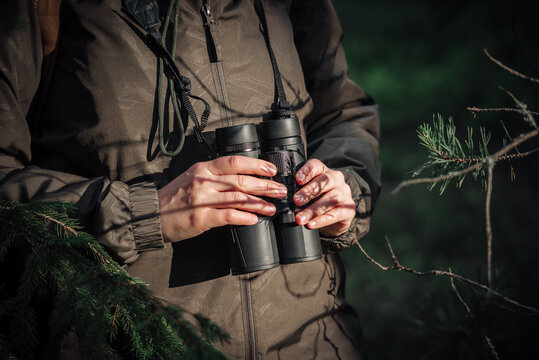 A girl in green jacket stands in the forest on a sunny day and holds binoculars in his hands.