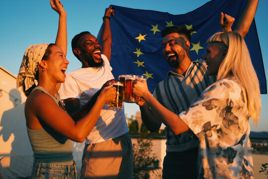 Happy friends enjoying a rooftop party, toasting with beer while celebrating europe day with european union flag