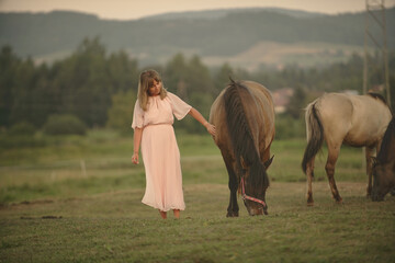  a woman in a pink dress standing near a horse in a grassy field with mountains in the background.