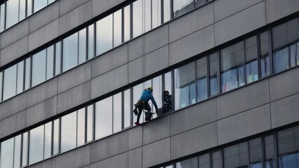 An industrial climber washes windows on the facade of a modern office building. A worker in protective gear and a helmet performs high-altitude work while hanging on a rope system. 