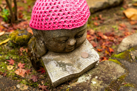 Jizo Statue with Pink Knitted Hat Resting on Book in Japanese Temple GardenJizo Statue with Pink Knitted Hat Resting on Book in Japanese Temple Garden