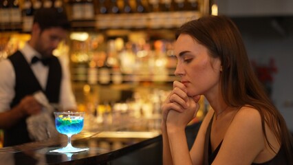 Sitting beautiful woman guest feeling bored waiting friends or couple with drinking mixed vodka in blue hawaii cocktail at luxurious counter bar for Friday meeting on bartender background. Vinosity.