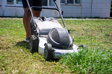 Unrecognizable man moves lawnmower across lawn to mow long grass in garden