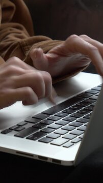 Vertical video of close-up female hands typing on laptop keyboard with light streaming through window