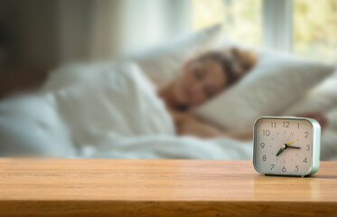Alarm clock on wooden table with sleeping woman in cozy bedroom bathed in natural morning light – morning routine and fresh start of the day. Copy space for product.