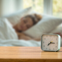 Alarm clock on wooden table with sleeping woman in cozy bedroom bathed in natural morning light – morning routine and fresh start of the day. Copy space for product.