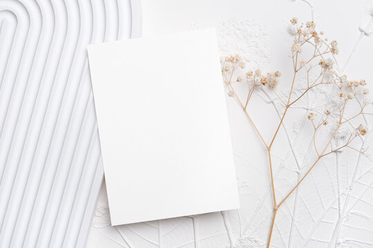 Blank white wedding invitation card mockup with delicate dried flowers on textured background, showcasing minimalistic design elements and soft natural aesthetics for creative projects