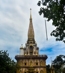 Golden Buddhist temple of Wat Chalong with intricate carvings, pointed spire, and surrounding greenery under a cloudy sky.