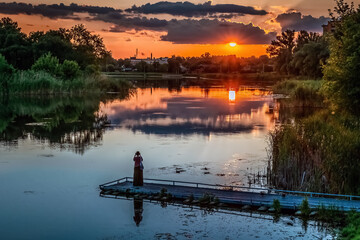 Woman standing on wooden pier watching dramatic sunset over Khorol River in Myrhorod, Poltava...